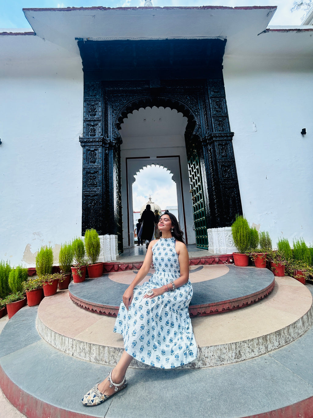 Woman in a floral dress sitting on a stone platform with decorative arches and plants in the background.