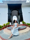 Woman in a floral dress sitting on a stone platform with decorative arches and plants in the background.