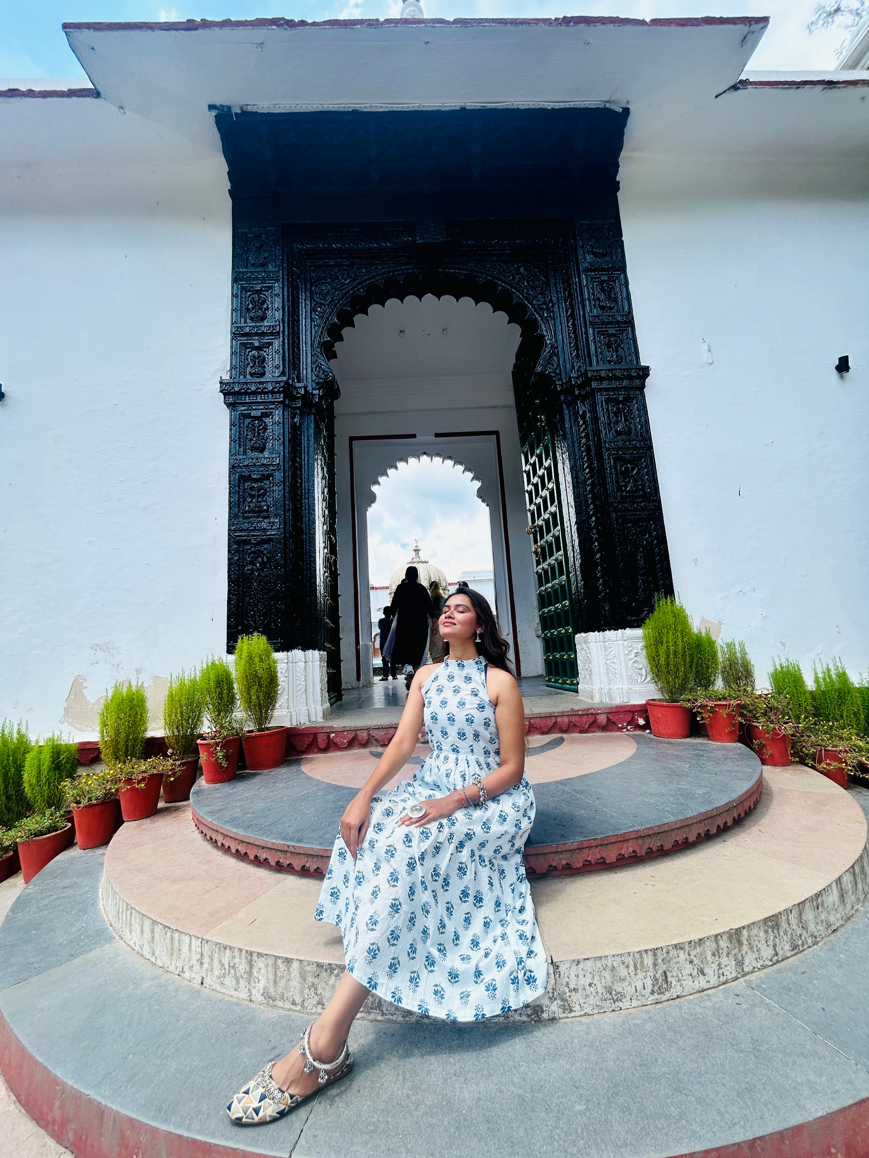 Woman in a floral dress sitting on a stone platform with decorative arches and plants in the background.