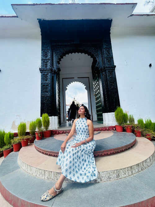 Woman in a floral dress sitting on a stone platform with decorative arches and plants in the background.