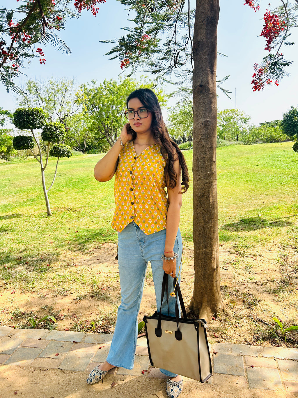 A woman stands outdoors in a park next to a tree, wearing a yellow sleeveless top with a small white pattern, light blue jeans, and holding a beige tote bag with black handles.