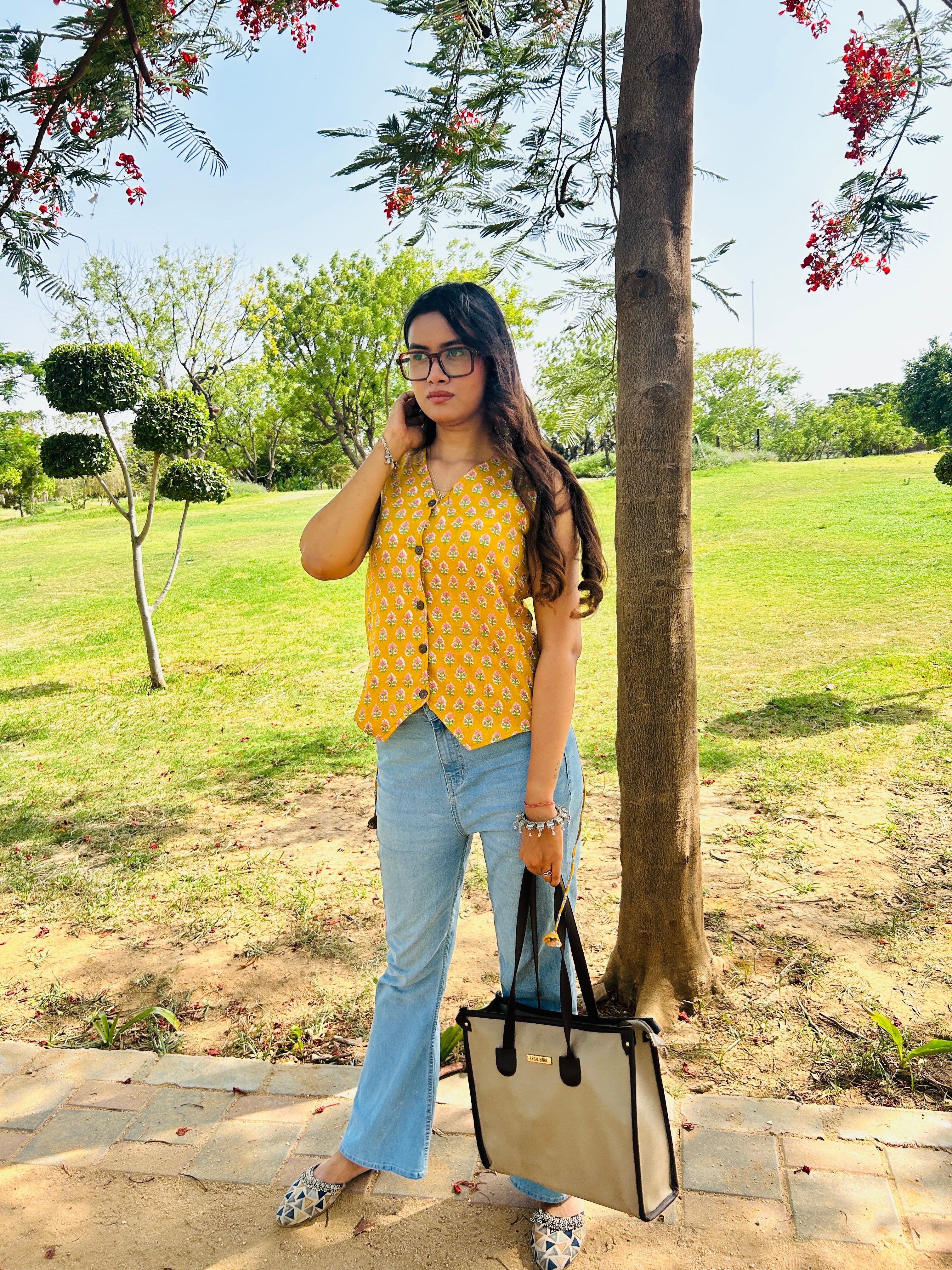 A woman stands outdoors in a park next to a tree, wearing a yellow sleeveless top with a small white pattern, light blue jeans, and holding a beige tote bag with black handles.