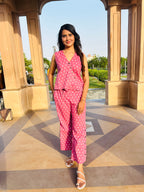 A woman modeling a pink sleeveless button-up top and matching wide-leg pants set with white polka dots, standing in an outdoor architectural setting with beige pillars and a patterned stone floor.