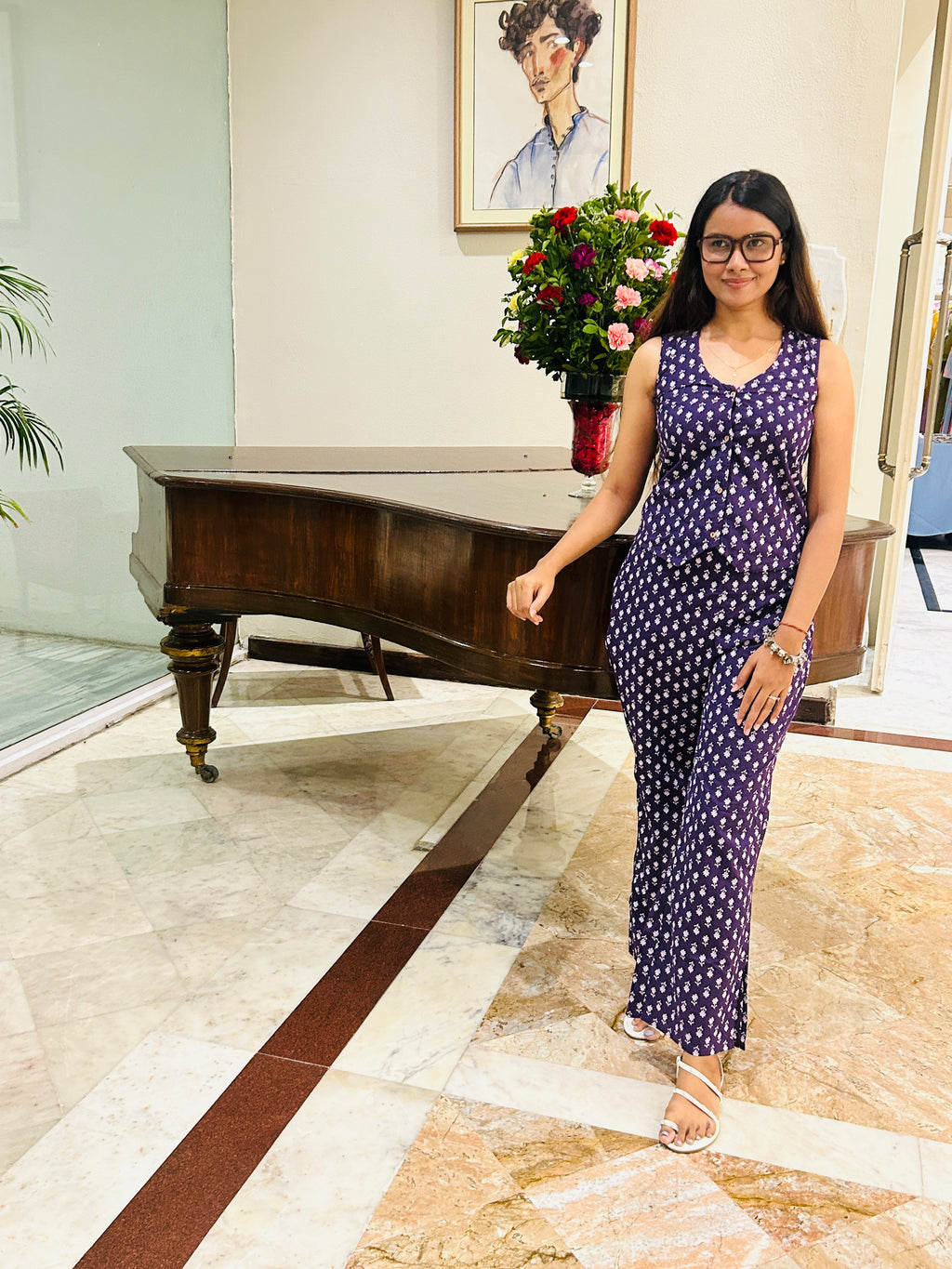 A woman wearing a sleeveless navy blue and white patterned jumpsuit stands in a lobby next to a grand piano.